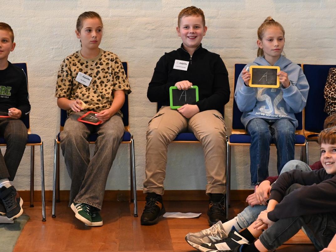 Jongens en meisjes die kandidaat zijn bij verkiezing kinderburgemeester zitten op een stoel met een krijtbordje met een antwoord